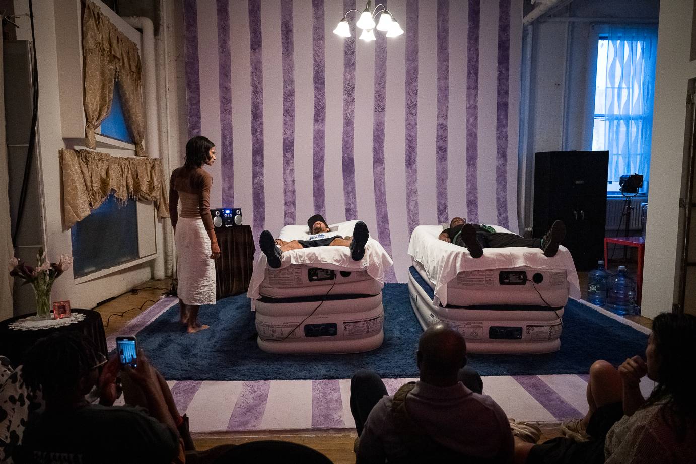 in a bedroom installlation with dark purple and lilac striped back wall and floor, are two twin beds, on which two audience members lie.  Ms tye z hovers about them.