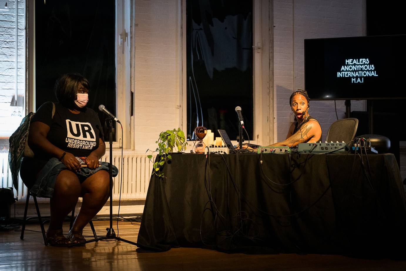 renata anakwe sits as a desk as a talkshow host interviewing a seated participatn who is a large black woman with a black t shirt  and denims skirt . renata looks out at the audience with her mouth wide open as if in suprise