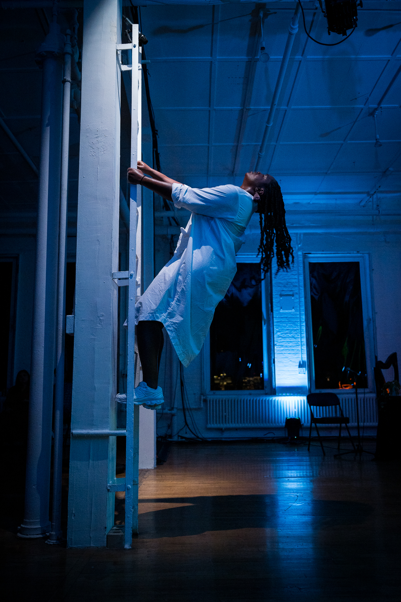 in blue light Ogemi Ude holds onto a ladder that is either an installation or part of the loft , in an oversized white shirt and sneakers she leans backward so her dread locks hang away from her body