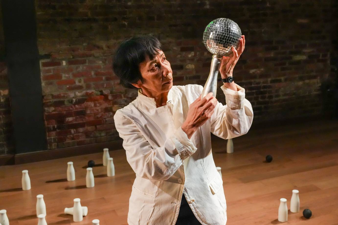 Woman in white jacket and short black hair holds up a metal orb to her left and stares at it. The background floor is littered with wooden, white milk bottles..