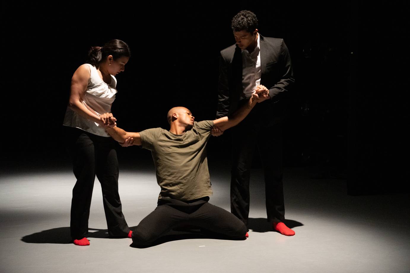 Abarakus Trio a woman and man in a suit tightly holde the arms of a man kneeling on the ground and looking pained on the floor