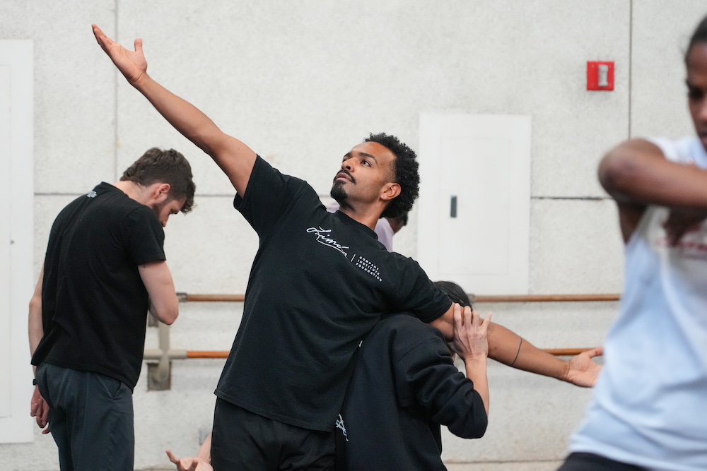 Eric Parra in a black t-shirt looks nobly out ward as he is about to be lifted by one of the other dancers in rehearsal -his arms are extended out horizontallly . he will be assuming the shape of a cross 