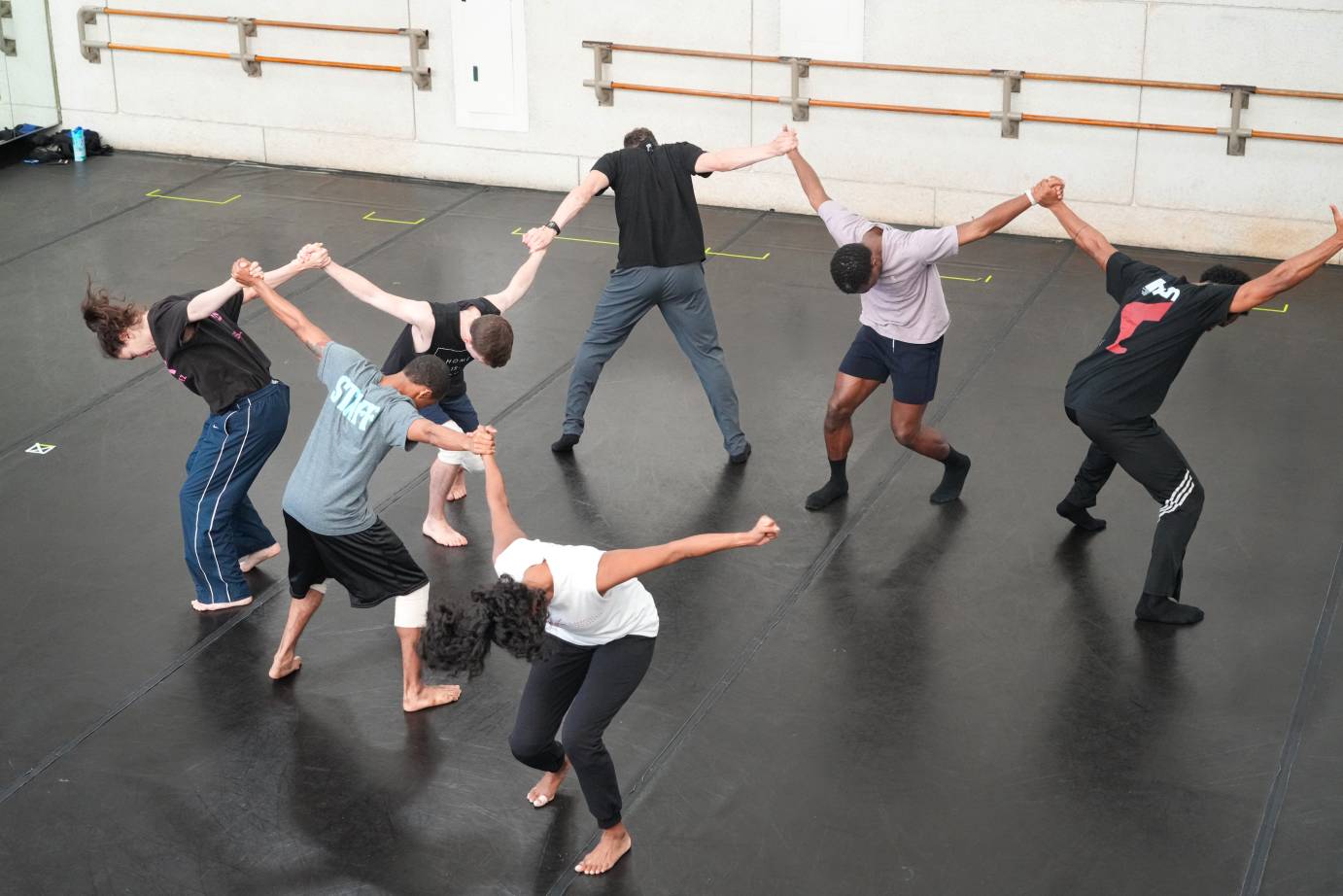 in the studio dancers holding hands in a semi circle bend forward facing different directions as their arms are flung upwards in a V shape