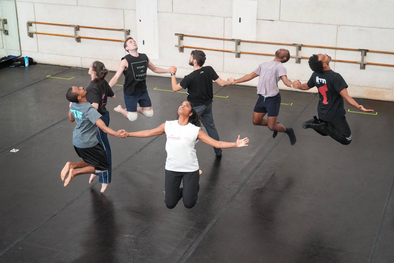 the photographer looks down on a semi circle of dancers in rehearsal as they hold hands and jump up folding their knees so their feet are behind them... their faces look upwards , except for one dancer in the center who stands as if to bring his friends down to earth