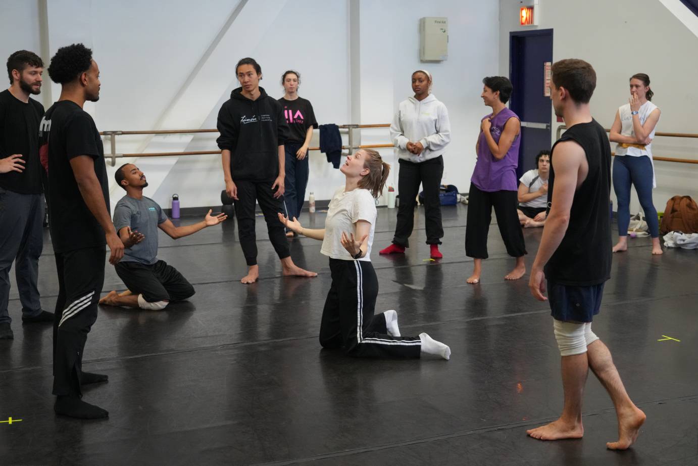 the rehearsal director kneels demonstrating to the company who surrounds her how to attack one of the movement in the dance 