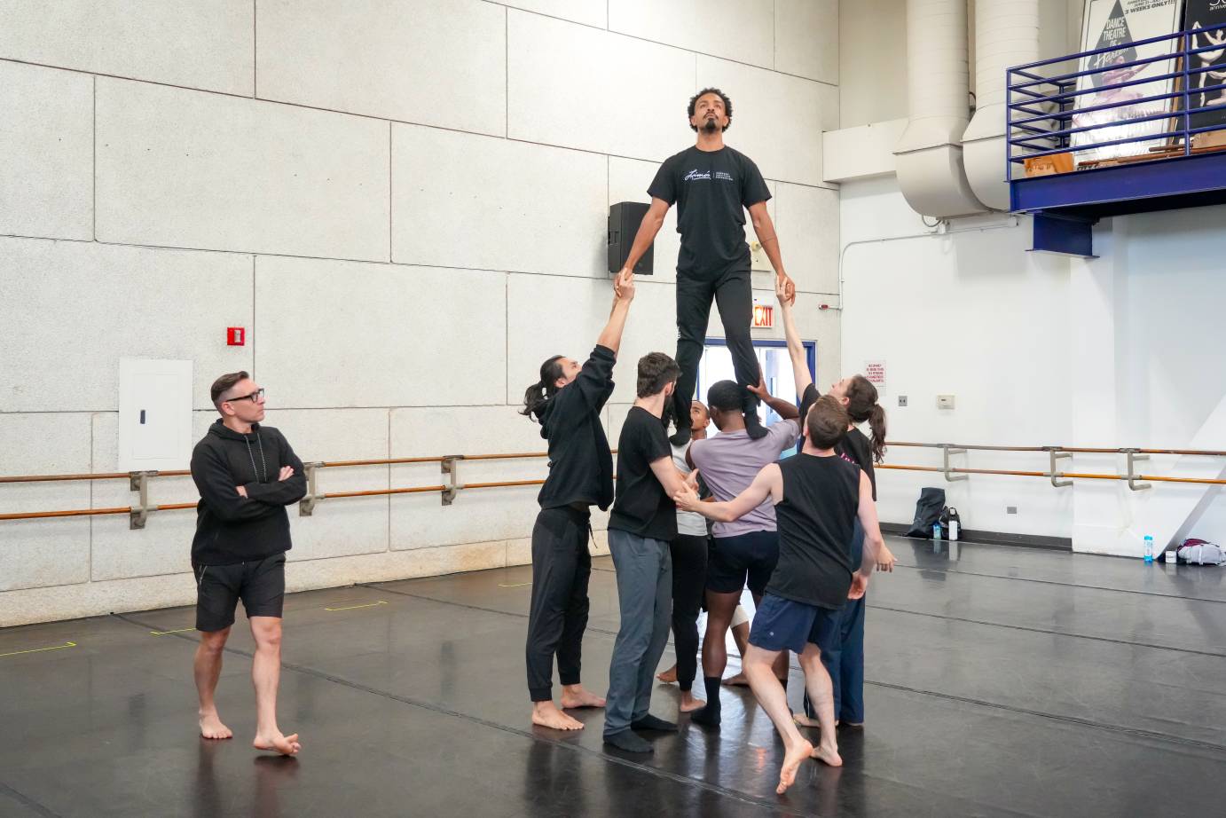 the company holds Eric the lead dancer as he stands on top of their shoulders and prepares to walk down along their bodies, Dante Puleio analyzes