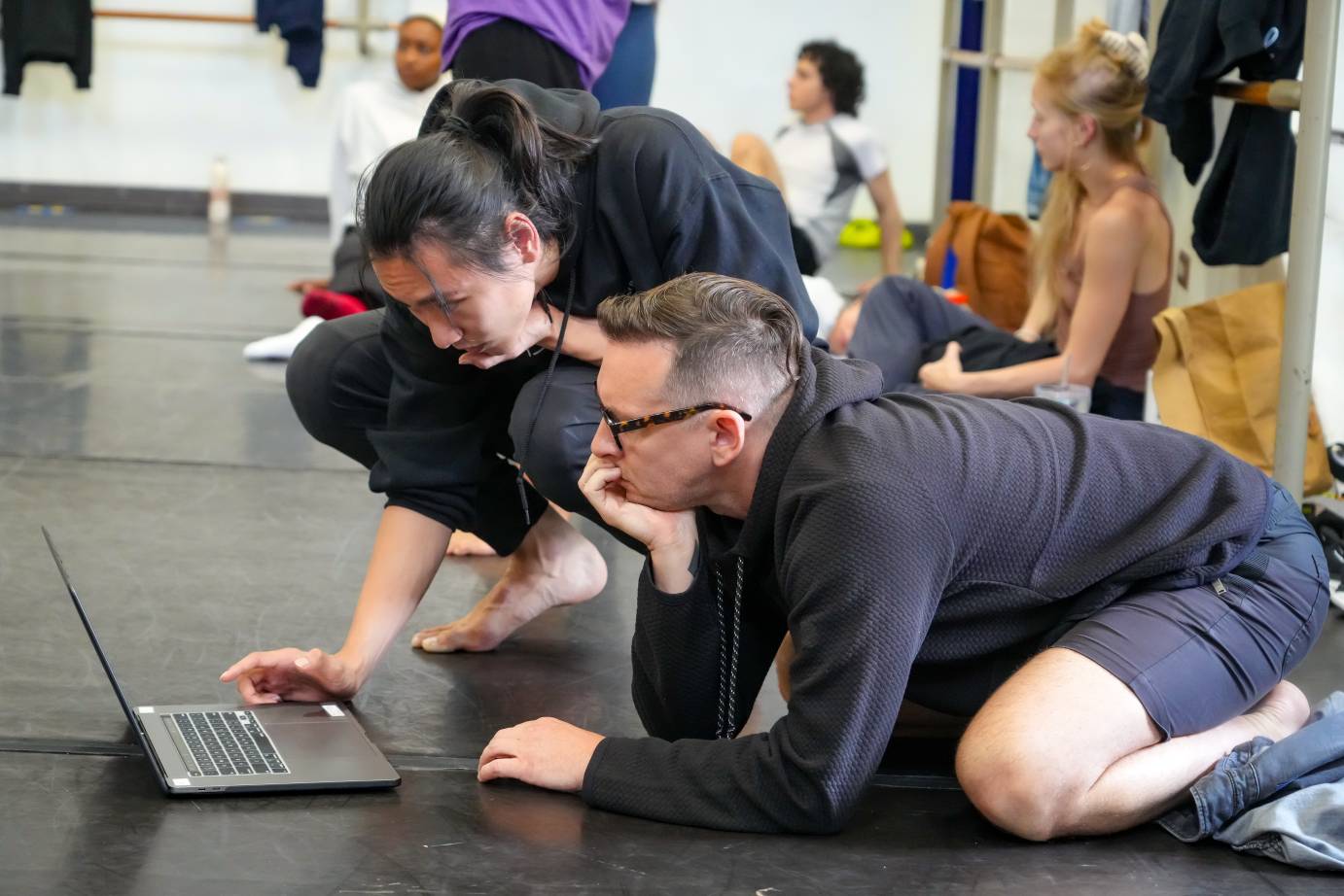 two dancers huddle in front of the computer to observe the original cast's work and compare it to their own