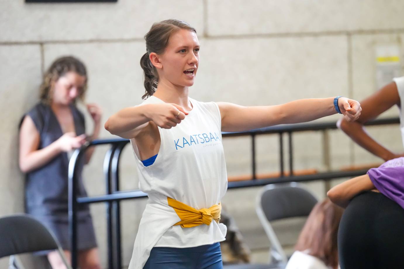 one of the women in a white sleeveless tshirt speaks to the group with her arms in an open wide circleshe is making an observation about the dance movement