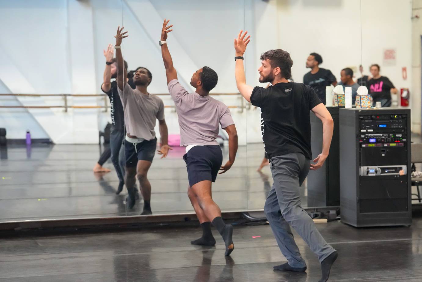 with the mirror to their side two dancers in rehearsal clothes lift their arms upward, one looks to the rehearsal director ( unseen) for notes