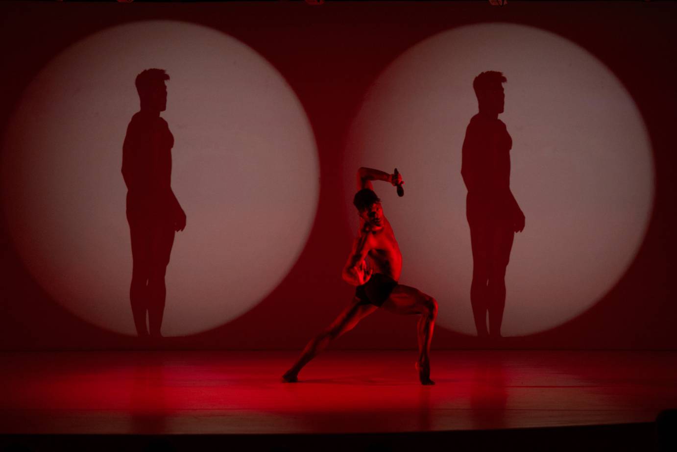 A man bathed in red lunges with his body side-lifting toward downstage. He poses between identical images of himself in lozenges in a film behind him.