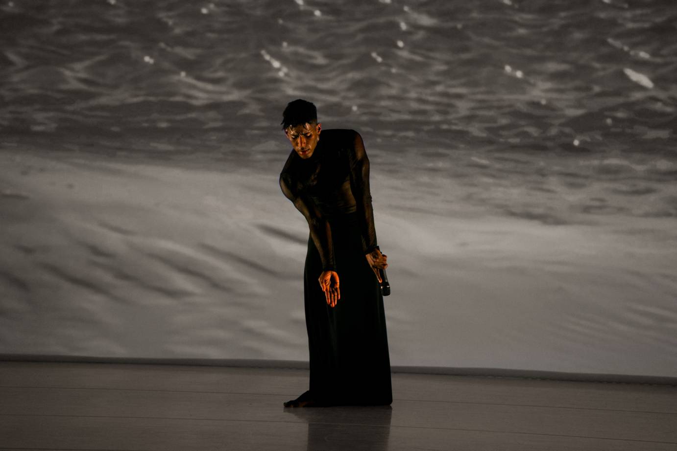 A man dressed in a long, black skirt and top stands with gaze downward and fingers snug together palms outward in front of a close up of the bubbling sea.