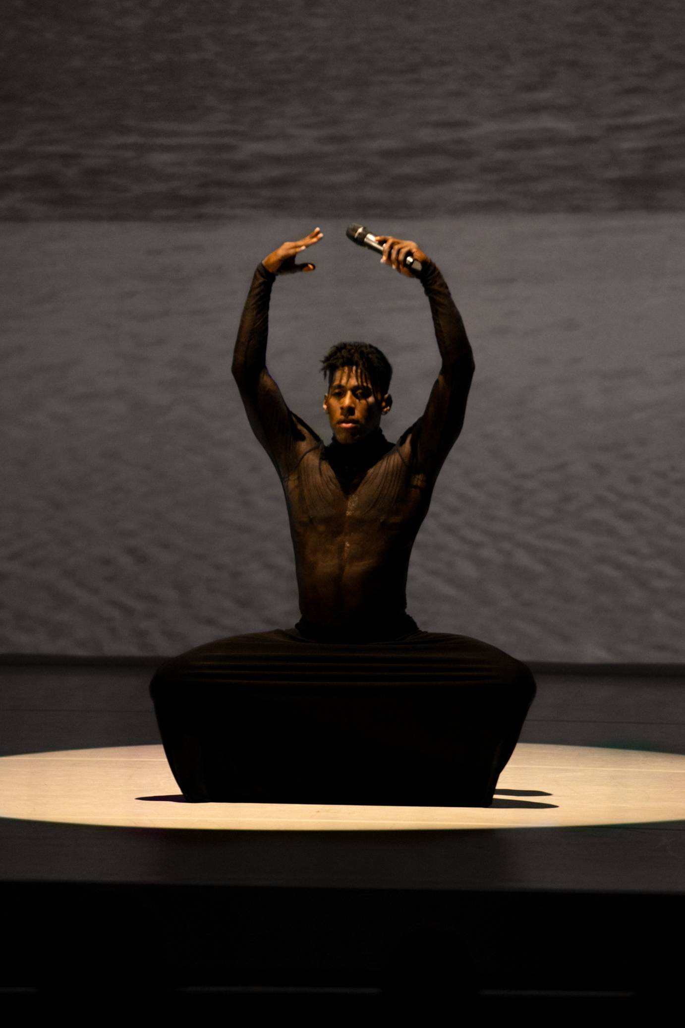 A man costumed in a black skirt and top is deeply plieing with arms overhead (one hand holding a microphone) in front of a film of water bubbles.