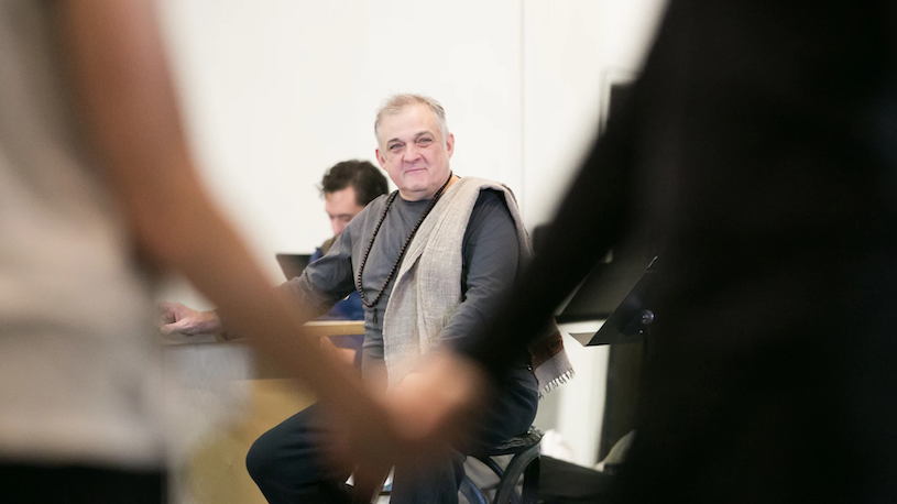 Mark Morris sitting down holding on to the barre looking at dancers in a studio rehearsal, he wears a black long sleeved t shirt with a tan scarf around one shoulder 