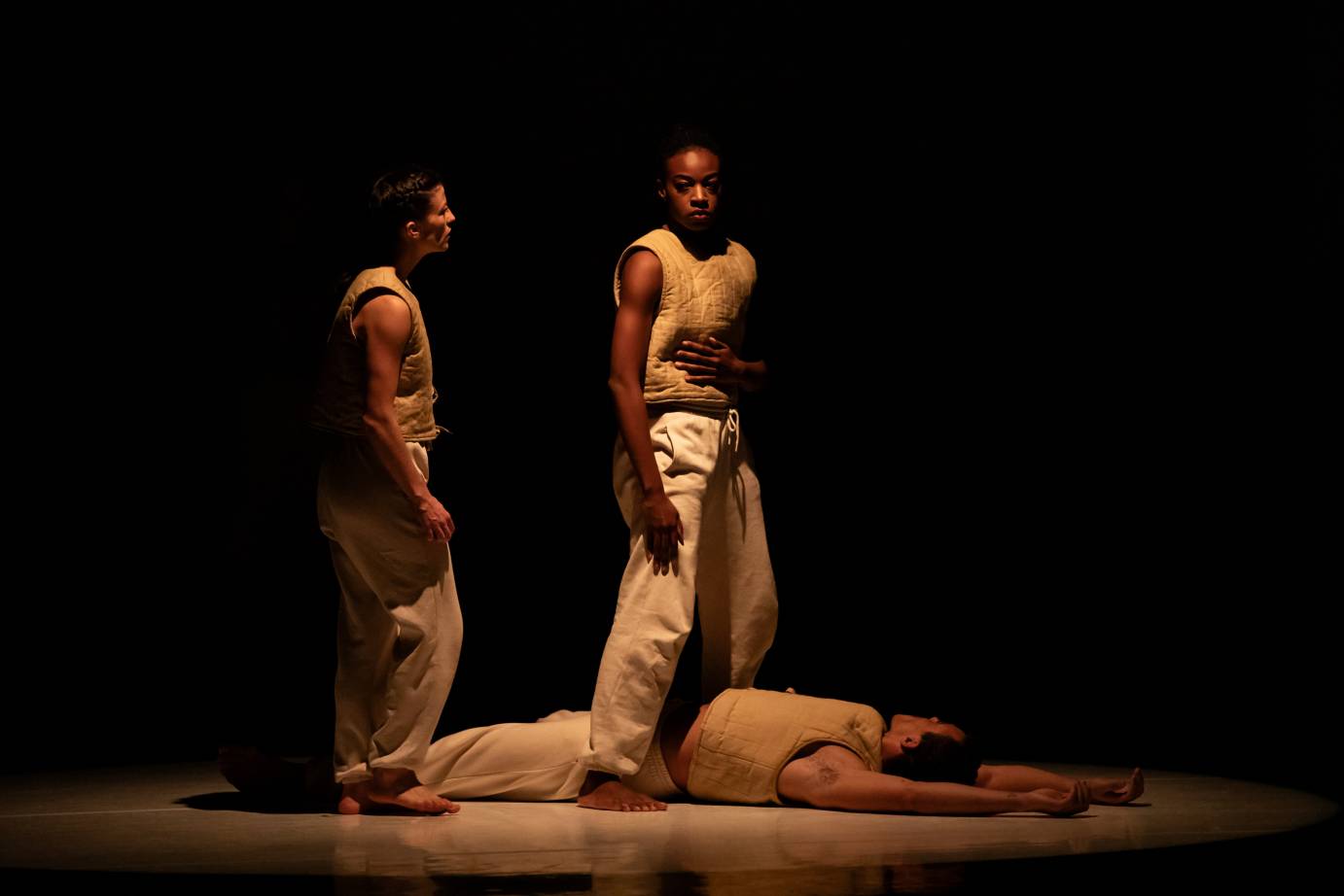 in darkness two dancers dressed in cream colored padded vests and sweat pants loom over another dancer ,dressed similarly, who, lying on the ground appears dead.