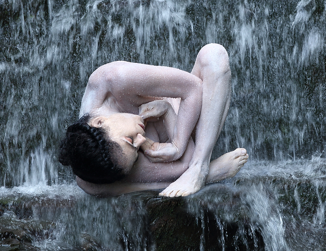 A woman's head, arms, and legs curled into her center perches on a waterfall ledge