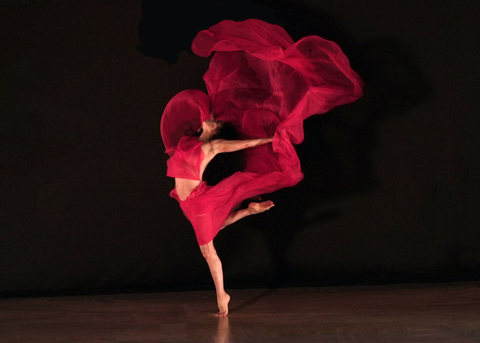 A woman on releve holding a swathe of red fabric that swirls around her body