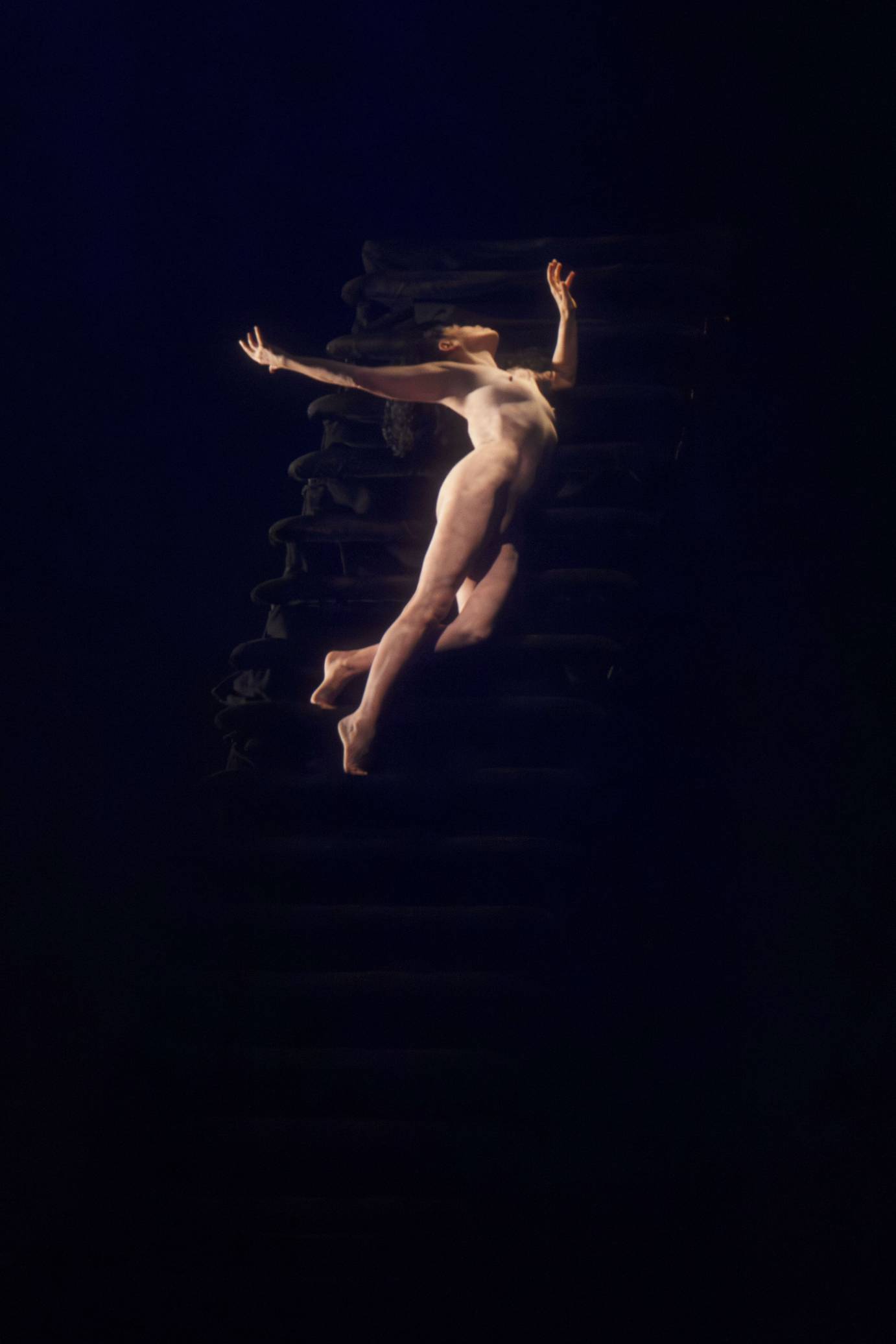 A nude woman on releve lifts her head and arms upward.