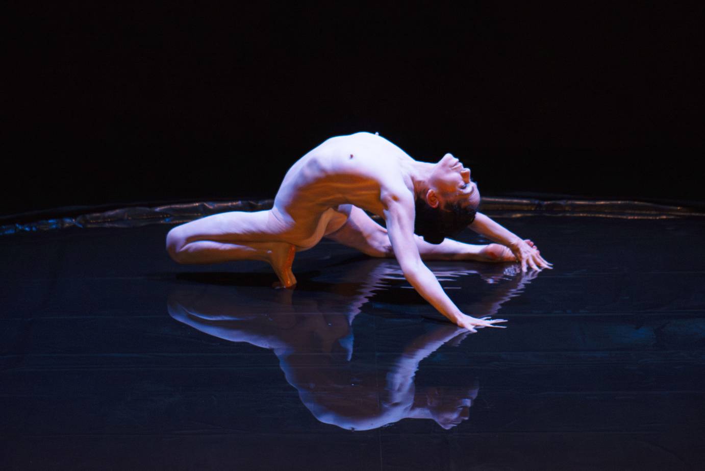 A nude woman kneeling in a pool of water arches backward