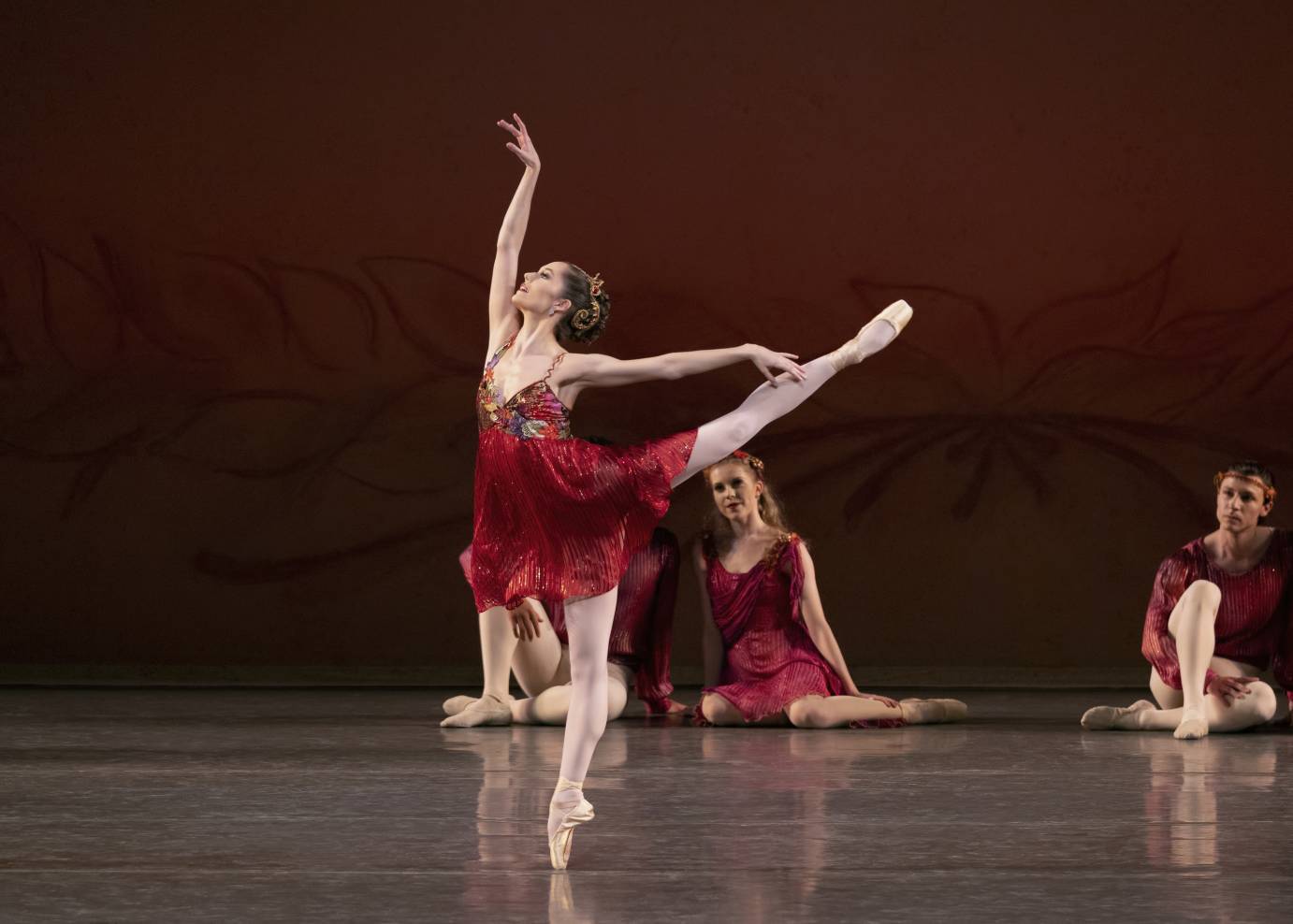 A ballerina in a mostly-red-inspired bodice and draping red skirt is on pointe in arabesque in front of three seated dancers.