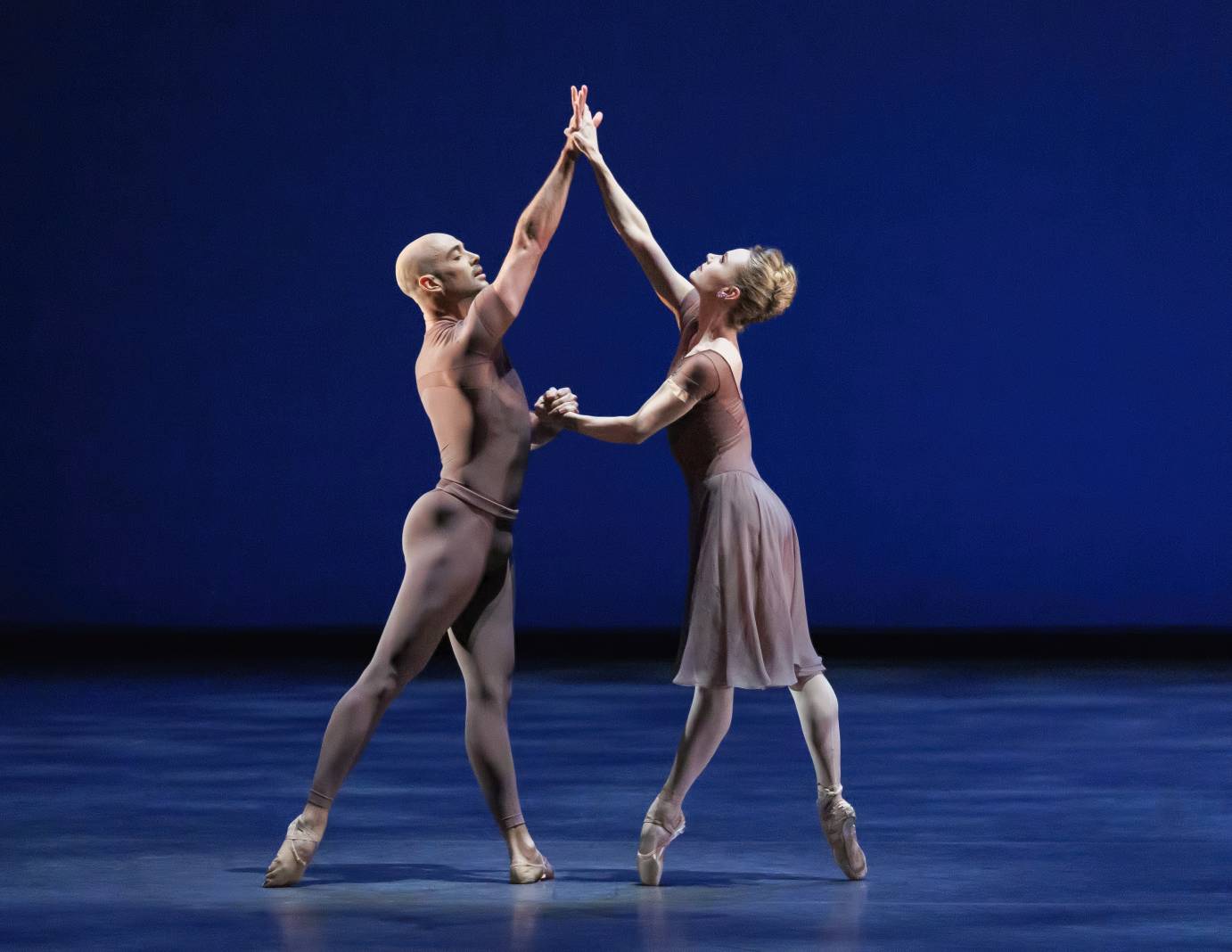 A man and a woman in sleek light aubergine-colored costumes stand on point clasping their opposite hands with the other as their opposite arms are raised above palms pressing.