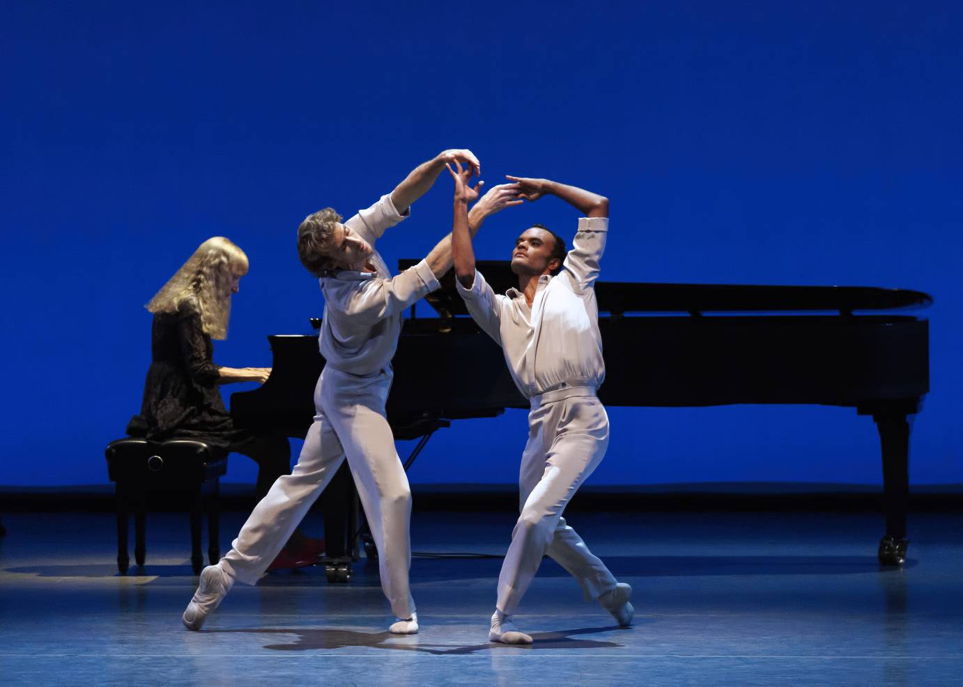 Two men dress in white shirts and slacks intertwine arms and hands extended above their heads as they dance in front of a grand piano played by a yellow-haired woman.