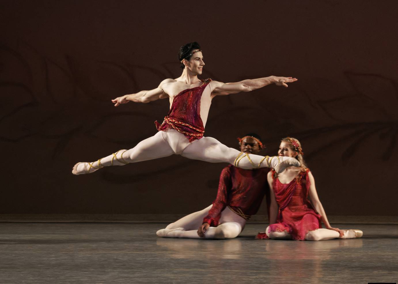 A black haired man leaps with horizontal legs in white tights and arms extended from a red topped torso. Two seated dancers watch him.