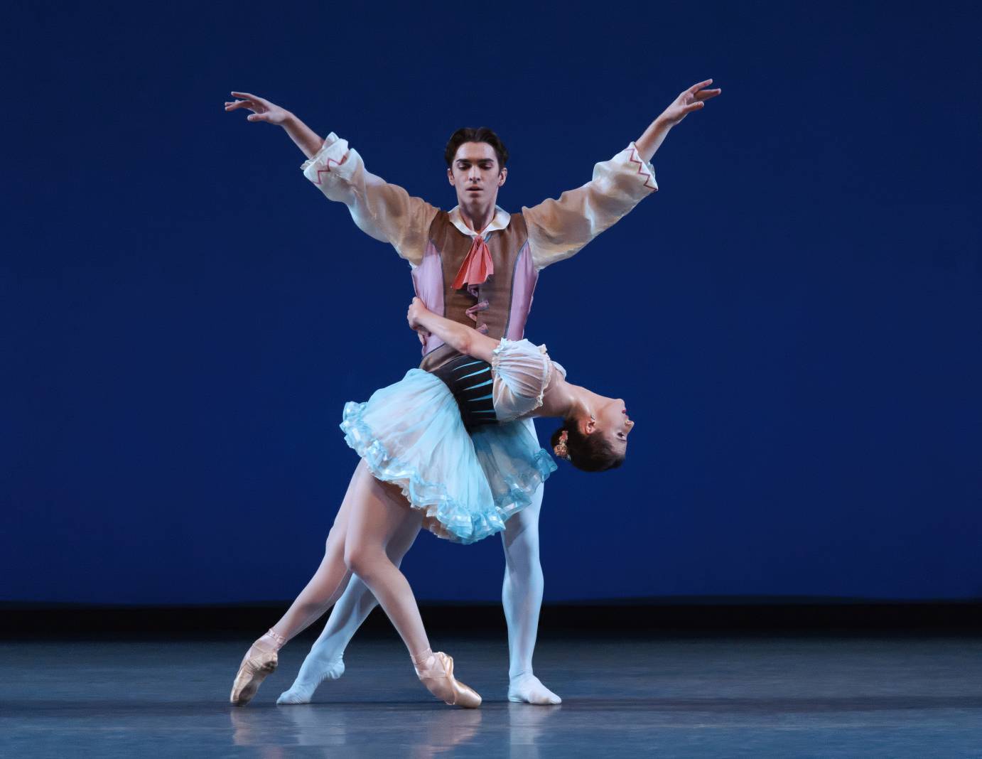 A ballerina arches backward holding herself up by encircling a man's waist. He is standing upright with his arms lengthened on the diagonal.