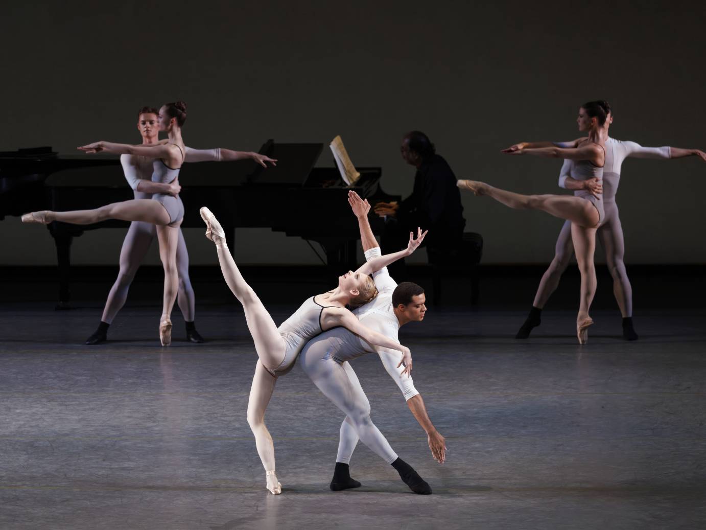 A central duet features a woman dressed in gray leotard and white tights with left leg extended upward rests on the back of a man with arms on diagonal - one arm toward the ground and the other skyward. Her leg and his arms are straight which is in contrast to her arms which look flung.