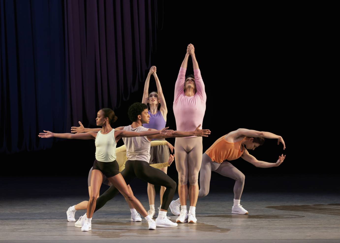 Five women and men dancers, costumed in many-colored tops, tights and shorts, and wearing white sneakers are grouped in long shapes, curved shapes and horizontal shapes on different facings.