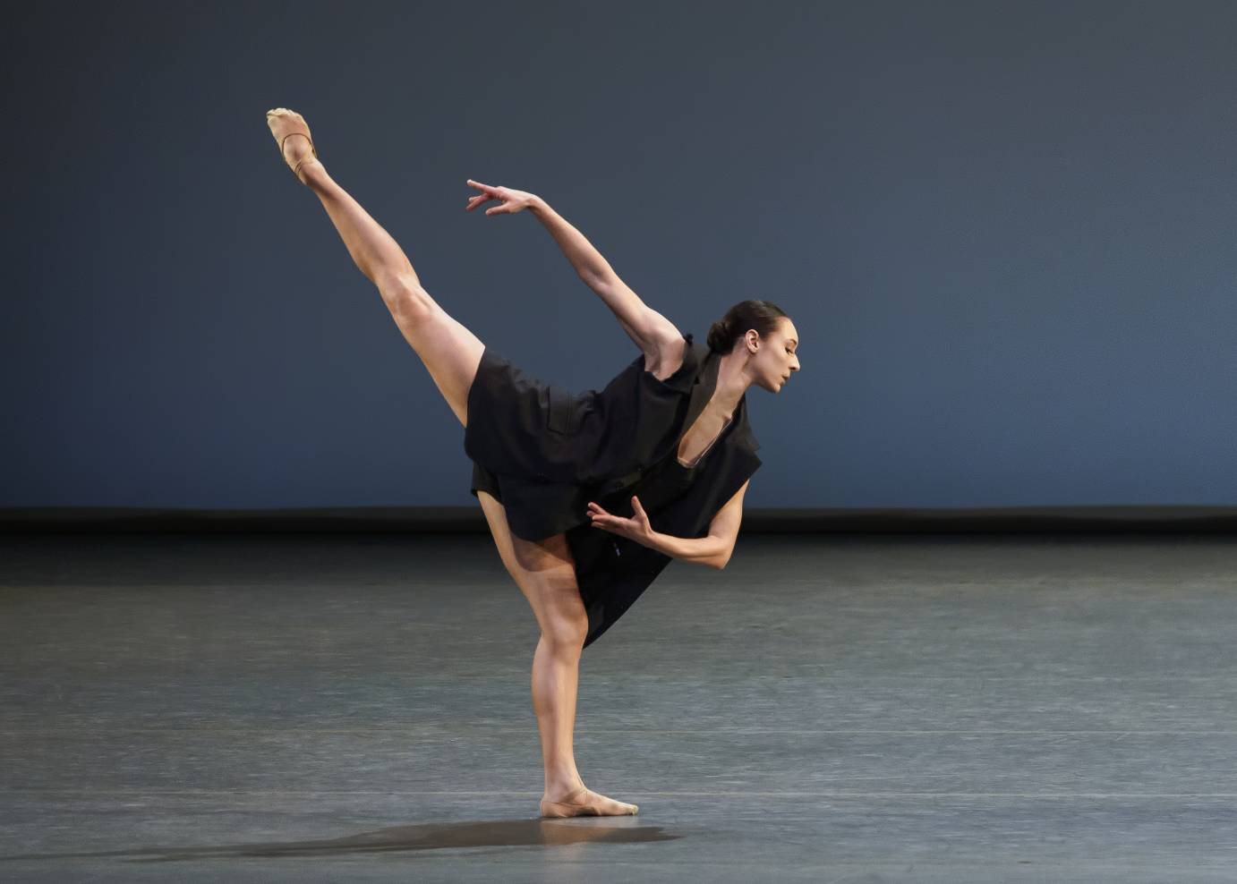 A solo woman dressed in a flouncy black dress and ballet slippers, and black hair in a low bun, looks down as her right leg and arm are upward.