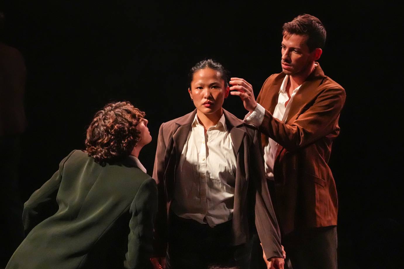 A trio of dancers dressed in brown and green suit jackers and white button down shirts are grouped with a woman in the center facing the auidenced while a man behind her reaches for her face and a woman below her intently looks at her face.