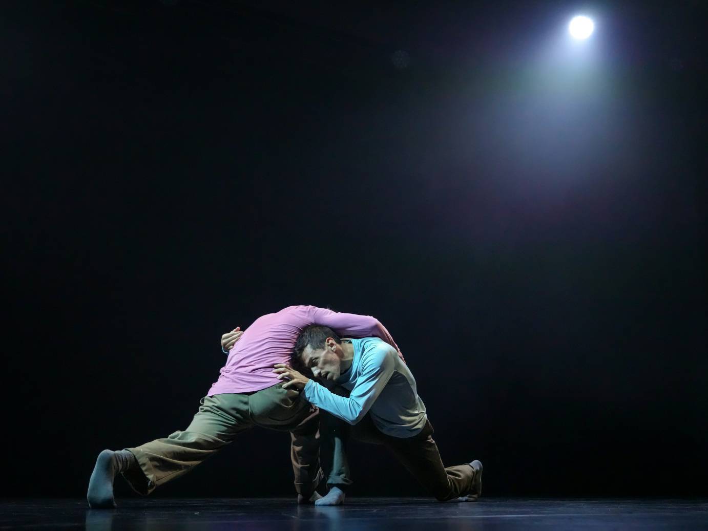 Two men grapple with one another: a pink shirted man drapes himself over a blue shirted man. Both lower bodies are lunging while their hands clutch the other.