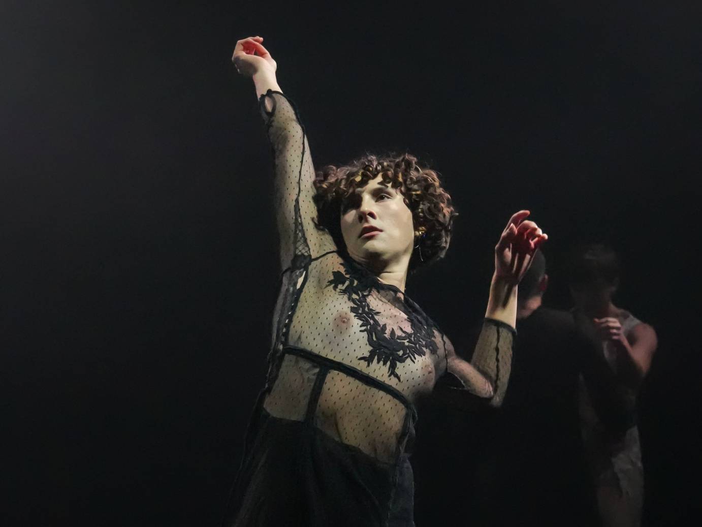 A curly-headed woman wearing a sheer black pointillist costume raises their right arm while the left is bent at the elbow.