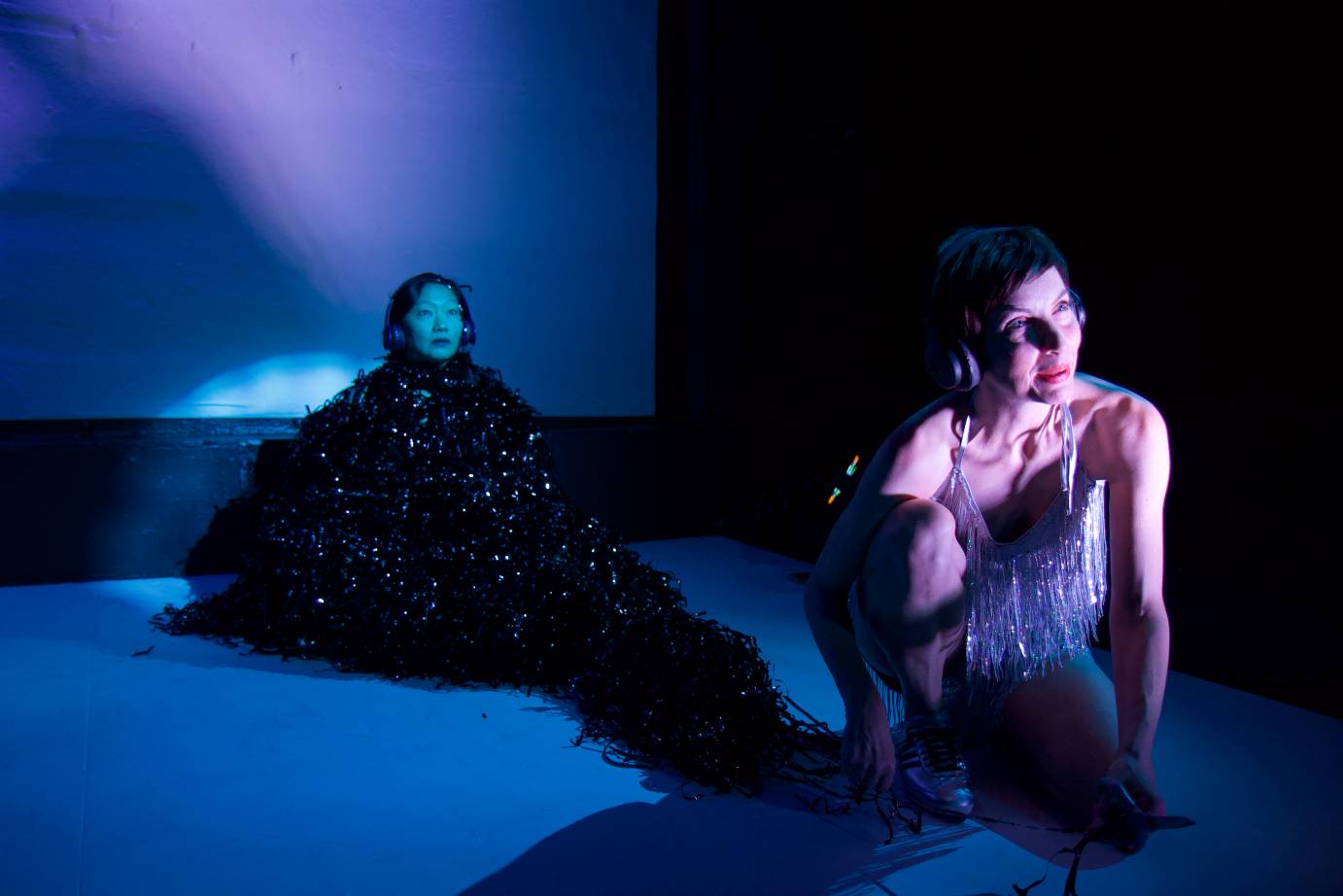 Two women sit on stage each looking on the diagonal. One woman is draped in mylar strips; the other is costumed in a fringed silver dress.