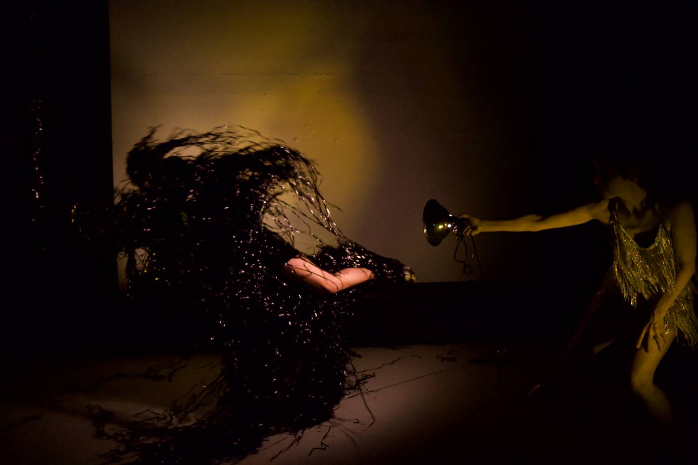 A woman draped in black mylar strips is spotlit by a woman in a silver fringe dress holding a spotlight.