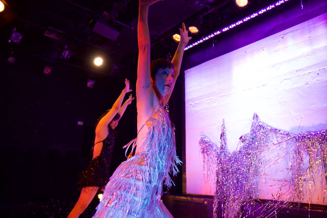 Two women stretch their arms high in front of the upstage screen draped by silver mylar strips.