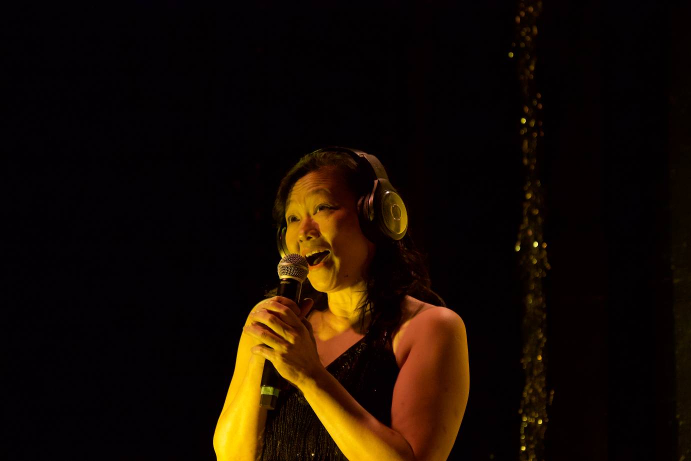 A woman wearing a black fringed dress speaks into a microphone that she is holding.