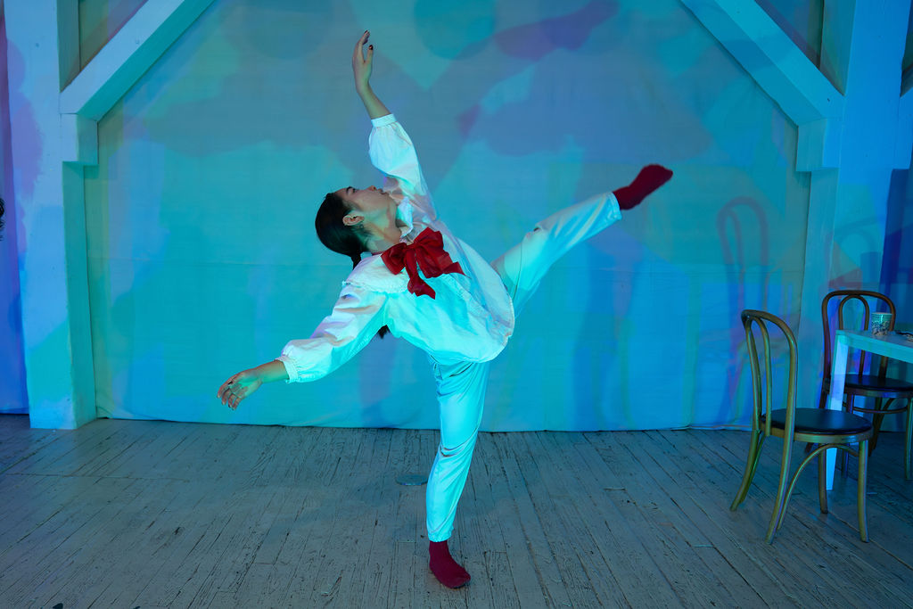 A dancer, dressed in white wearing red socks and a big red bow at her neck, lifts their left leg and left arm high.