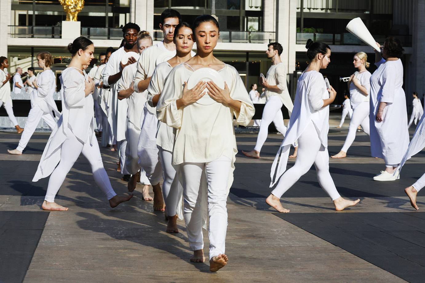 a line of dancers in white holding plates to their hearts come toward us as other dancers move in lines of other directions