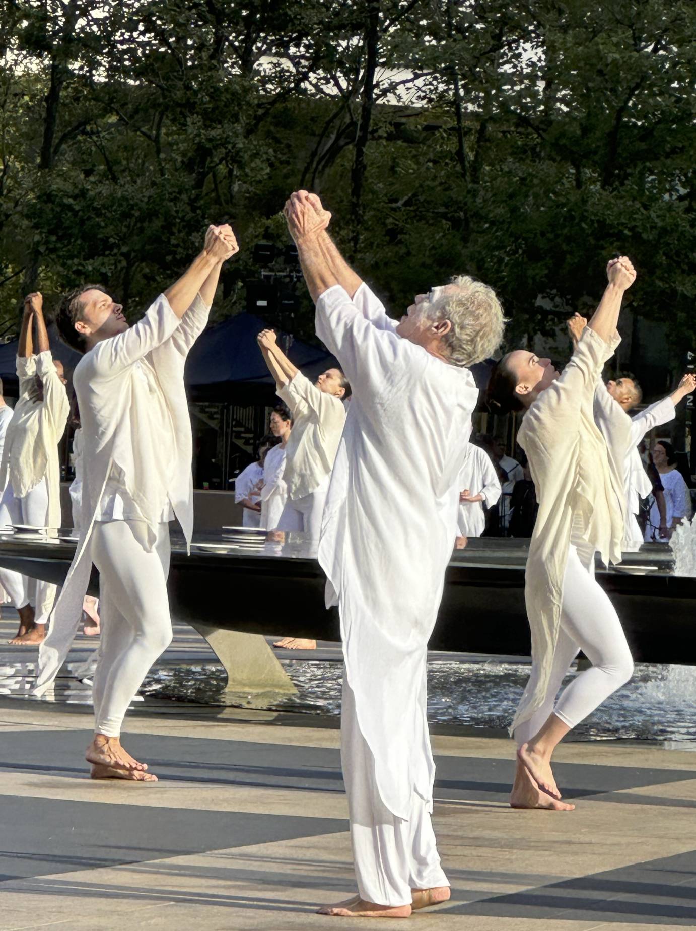 dancers in white performing a ritual around the fountain at lincoln center . they hold their hands and reach to the sky