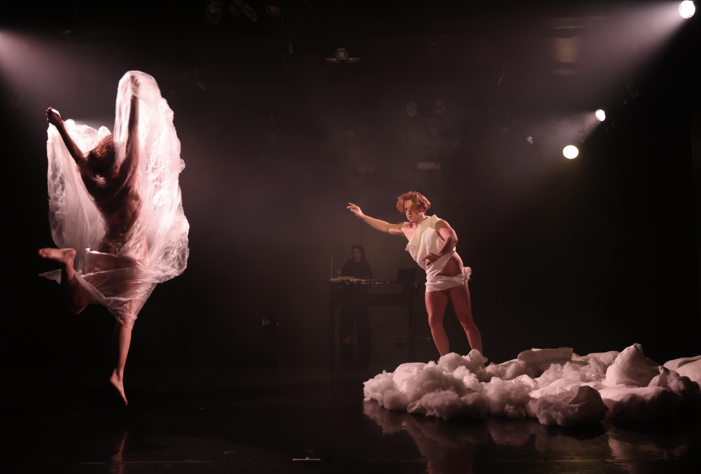 nude dancer jumping her back facing the audience surrounded by billowing plastic to her side a man wrapped in a white sheet barely covering his genitals and resembling a mini toga leans toward the billowing dancer standing on an island of pillows