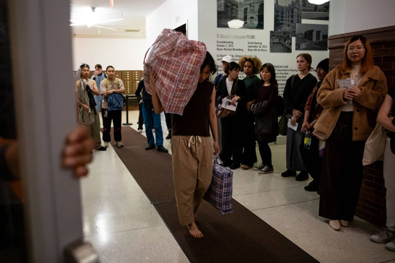 walking on a brown carpet a dancer in sweats holds a plastic suitcase on her shoulder as she moves in a line on a brown carpet. The audience is standing around her