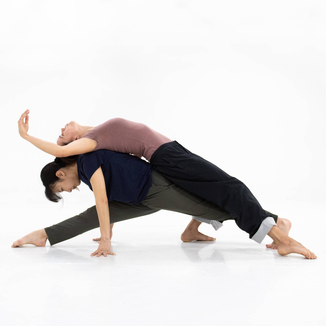 A female dancer drapes drapes her skyward-facing body over the body of another woman who is in a near split facing the floor.