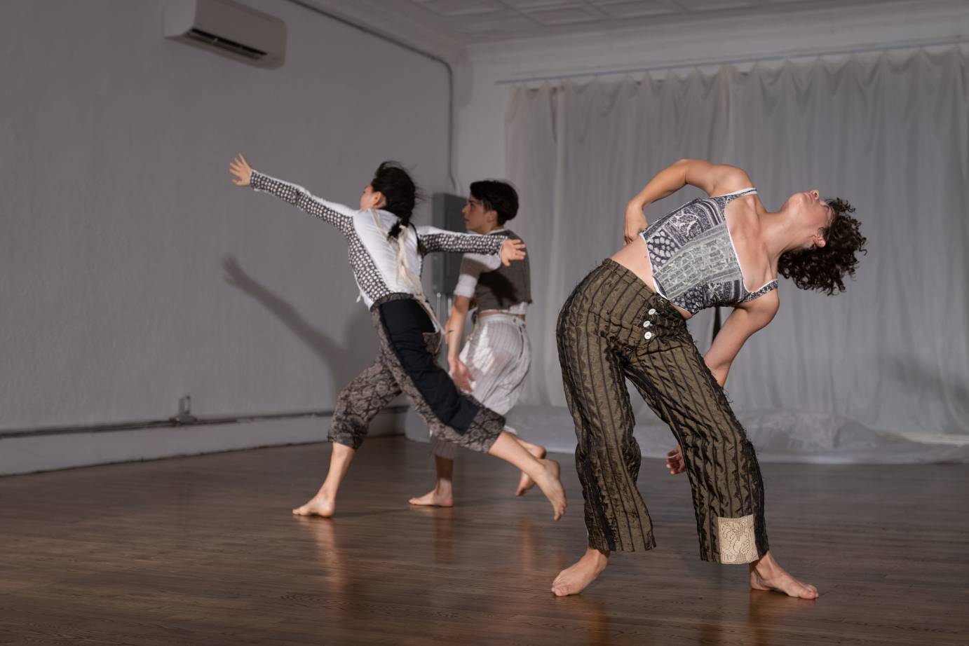 Three women dancers - one upstage in a standing contraction in profile facing stageright, one striding with wide legs and long arms in opposition in profile facing stage right and the third upstage in a standing parallel plie facing the ceiling