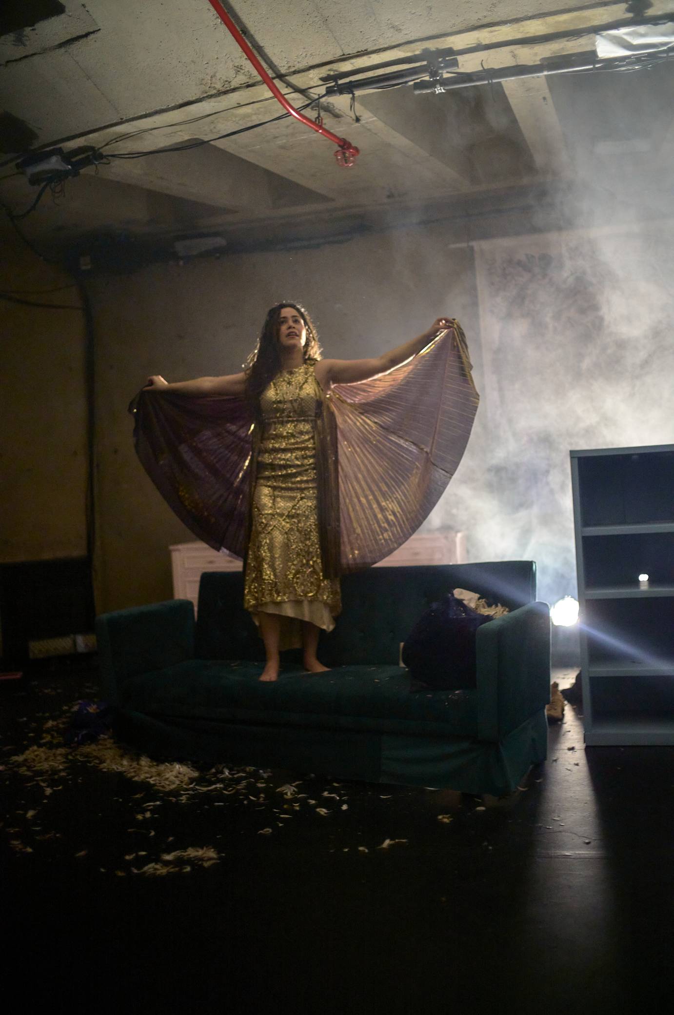 the woman with wet loose hair stands on a couch wearing a glitterin gold outfit with wing extensions that she holds spread out as if an angel. 