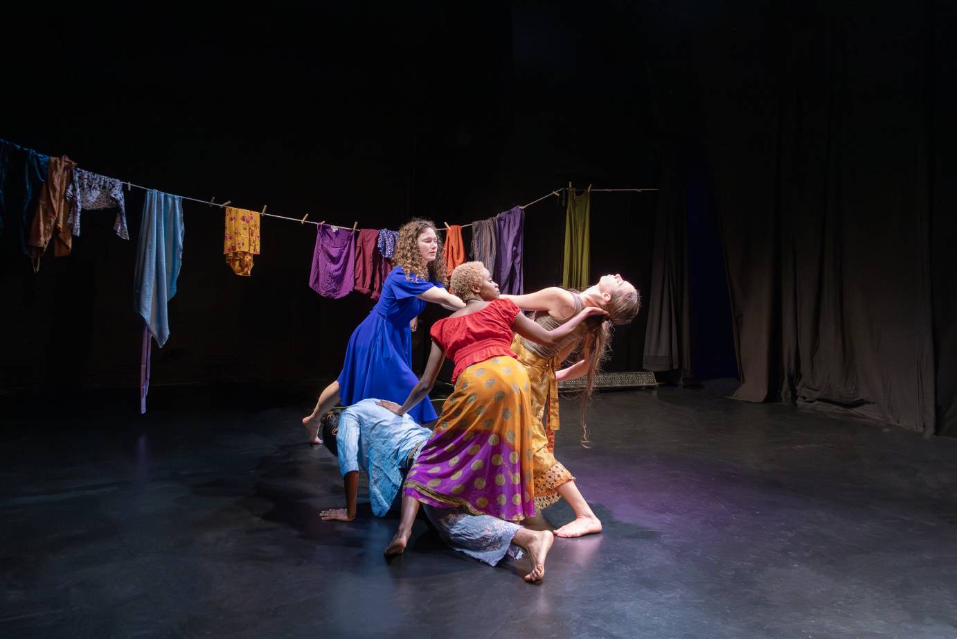 a tableau of women who dressed colorfully as they support one of their group who is leaning backwards away from a clothesline that hangs upstage