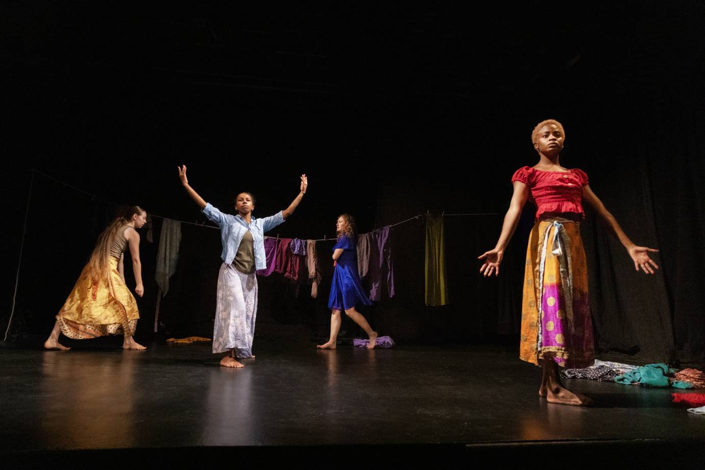 four women poised in shapes that indicate worship or contemplation stand before a clothesline filled with hanging laundry